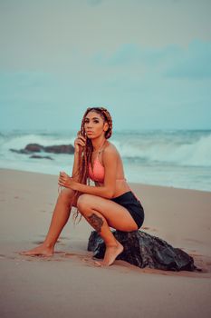 A woman with braided hair poses confidently on a beach, capturing the essence of summer fashion and ocean vibes.
