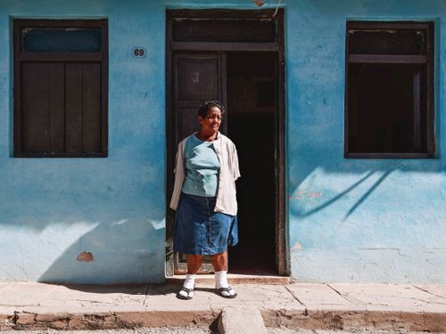 Free Senior woman in casual attire stands in doorway of rustic blue house. Stock Photo