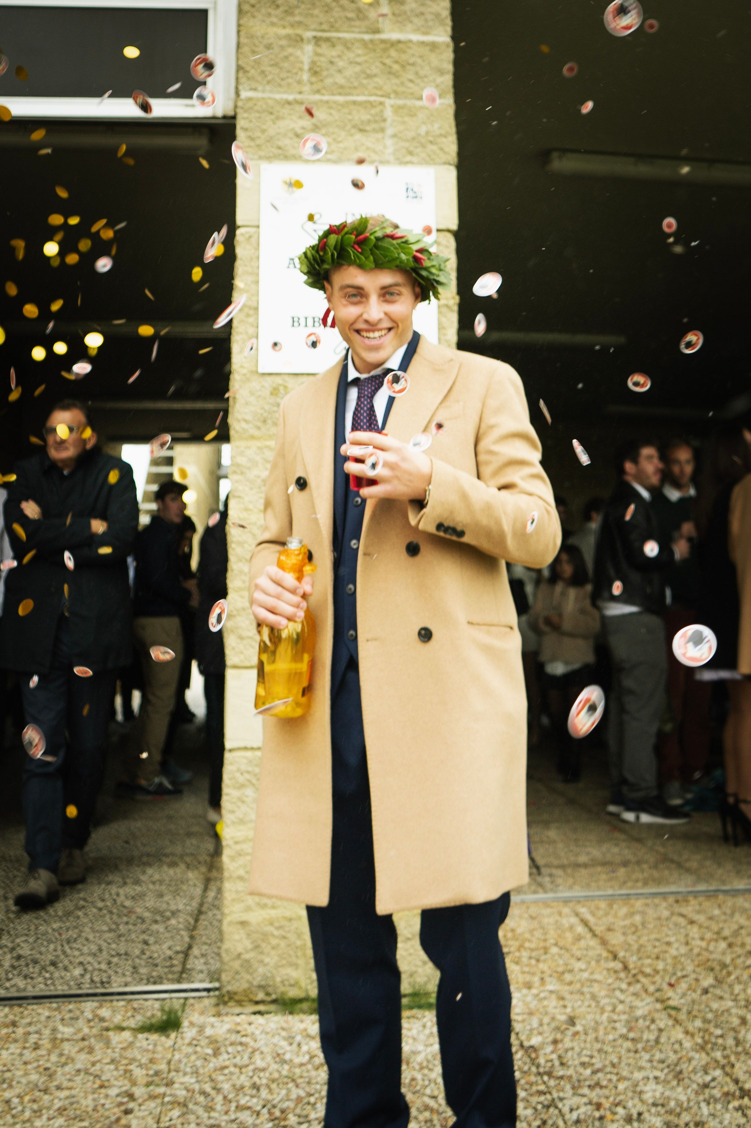 Smiling man in coat celebrates graduation day with champagne and confetti outdoors.