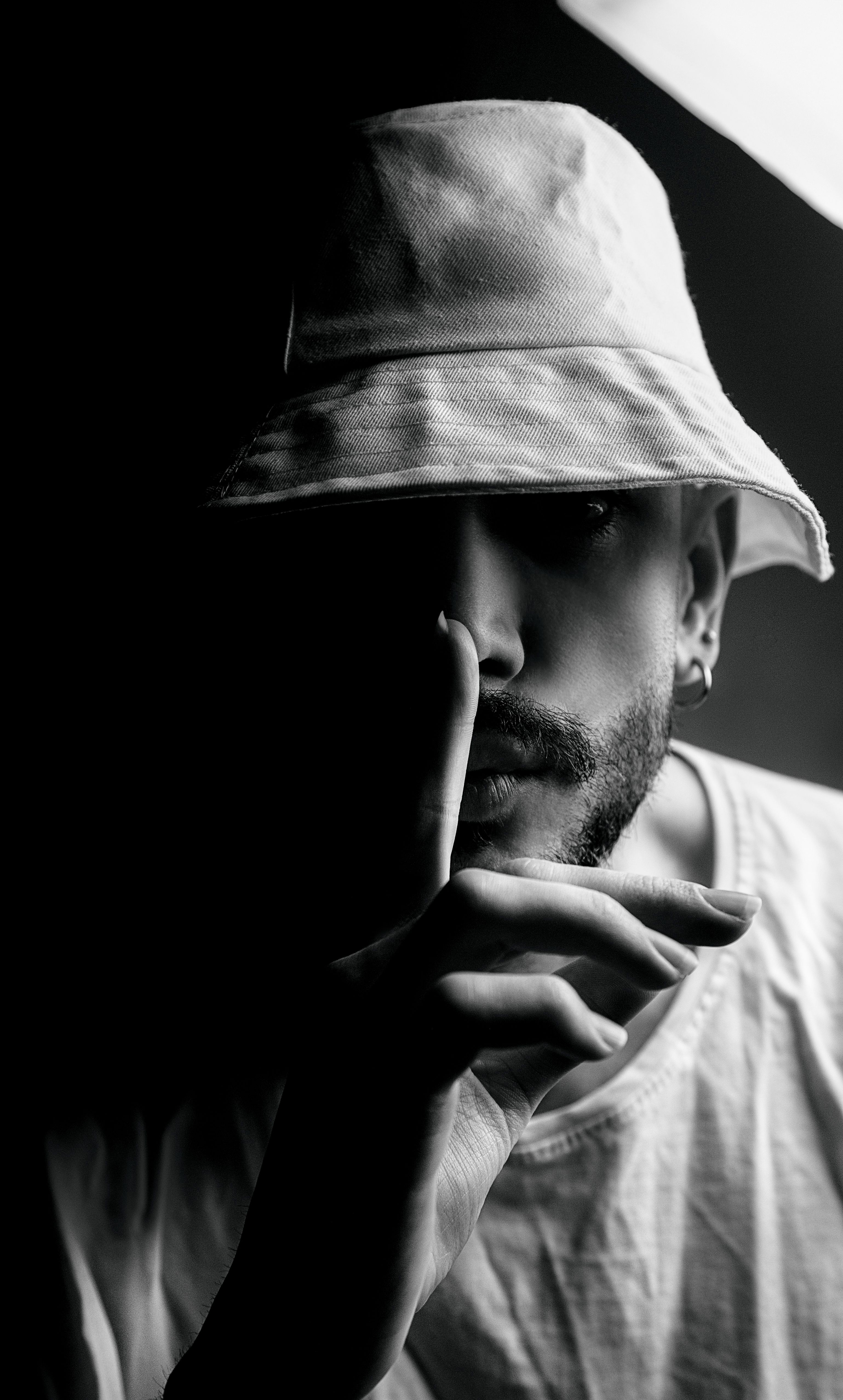 Black and white portrait of a man wearing a hat, with finger on lips in a shushing gesture. Dramatic lighting and shadows.