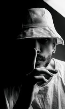 Black and white portrait of a man wearing a hat, with finger on lips in a shushing gesture. Dramatic lighting and shadows.