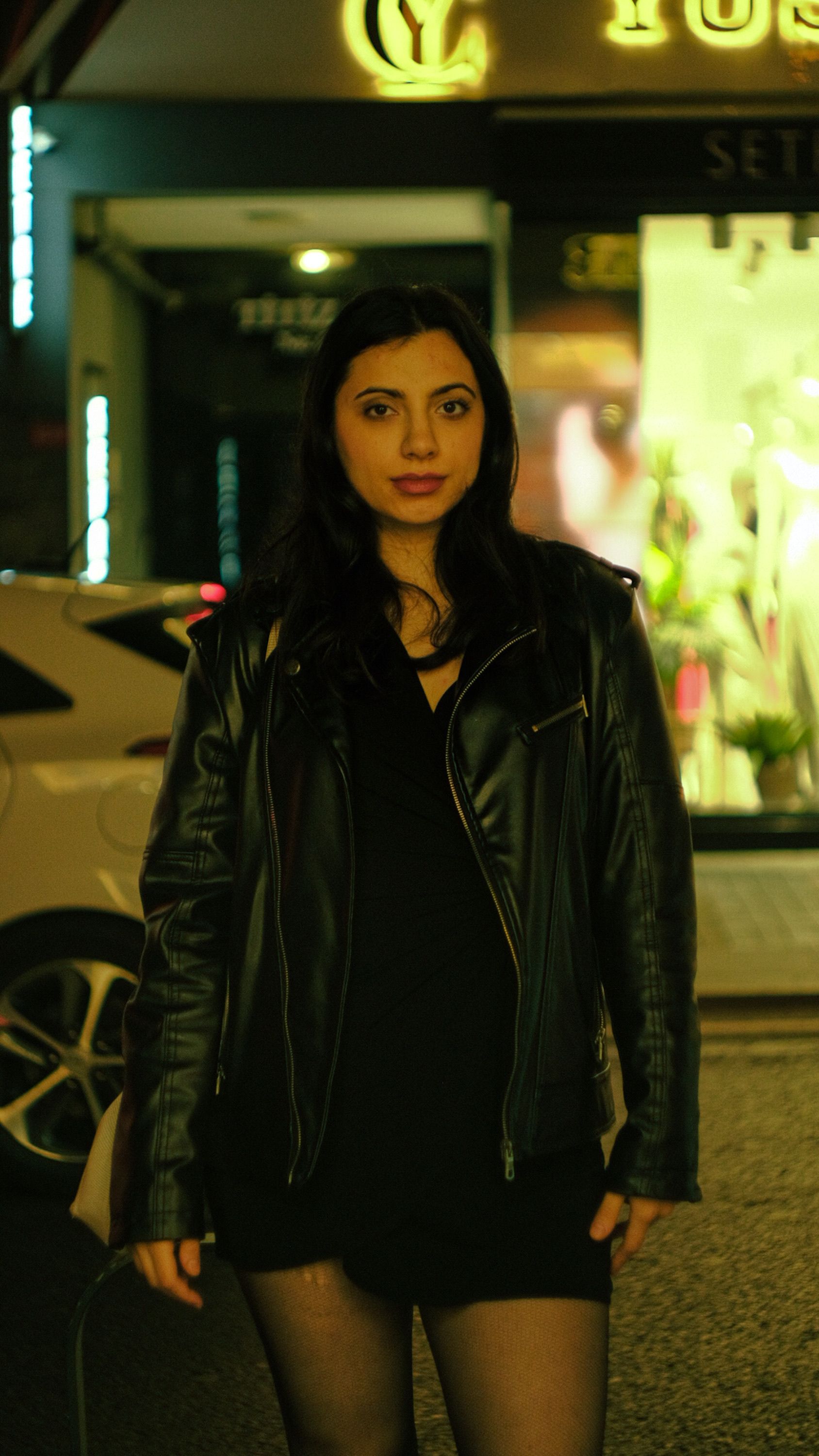 Stylish woman in black leather jacket posing on city street at night, exuding confidence and elegance.