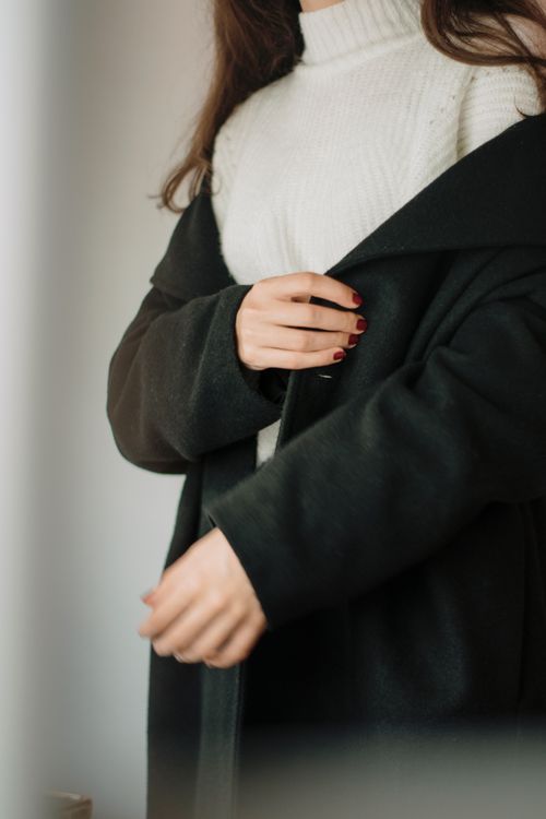 Free A fashionable woman adjusting her black coat over a white knit sweater indoors. Stock Photo