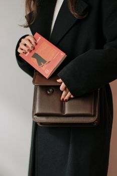 A stylish woman in an elegant coat holds a brown leather bag and a book. Fashion and sophistication captured.