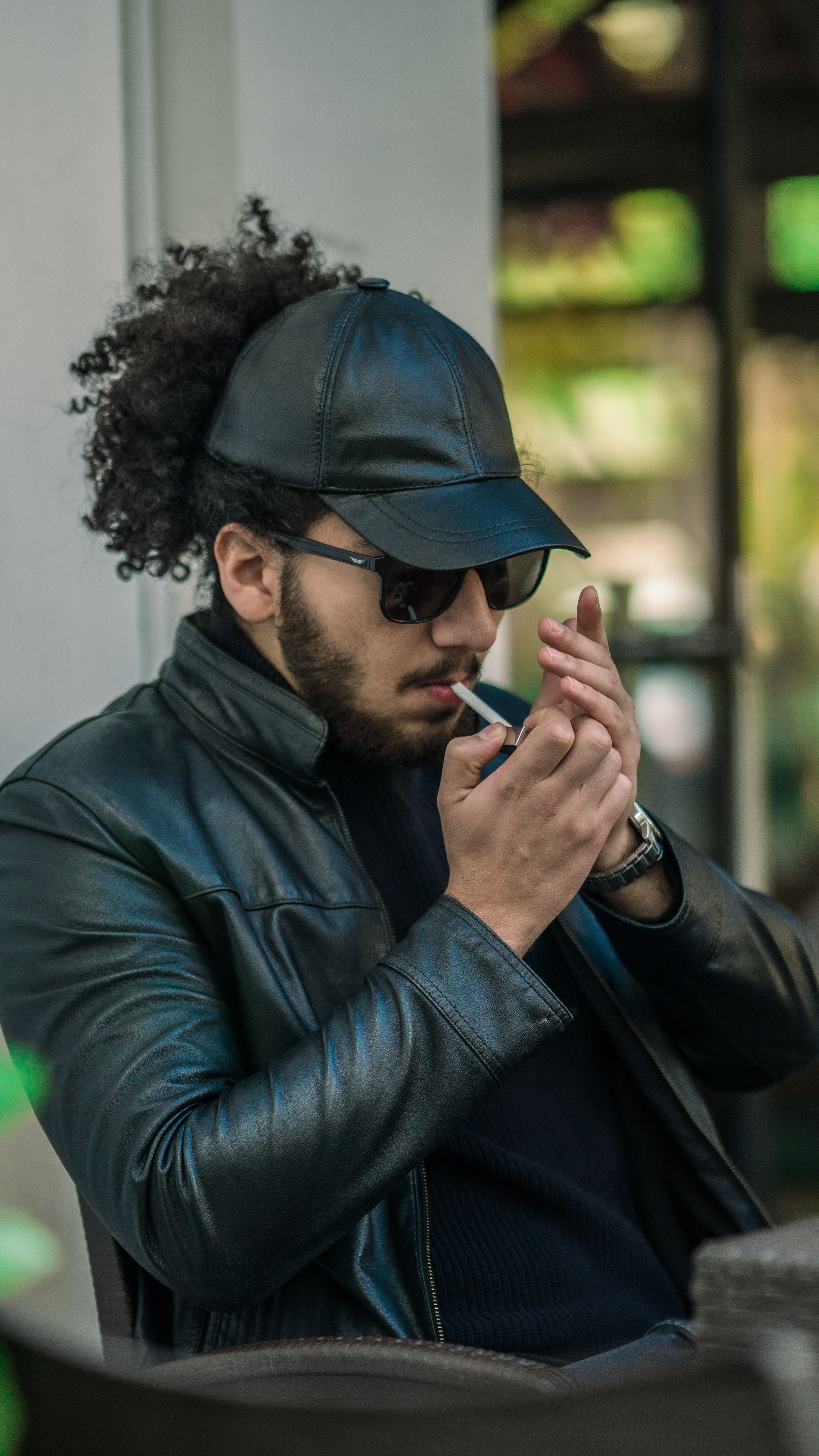 Stylish man in leather jacket with sunglasses lighting a cigarette outdoors.