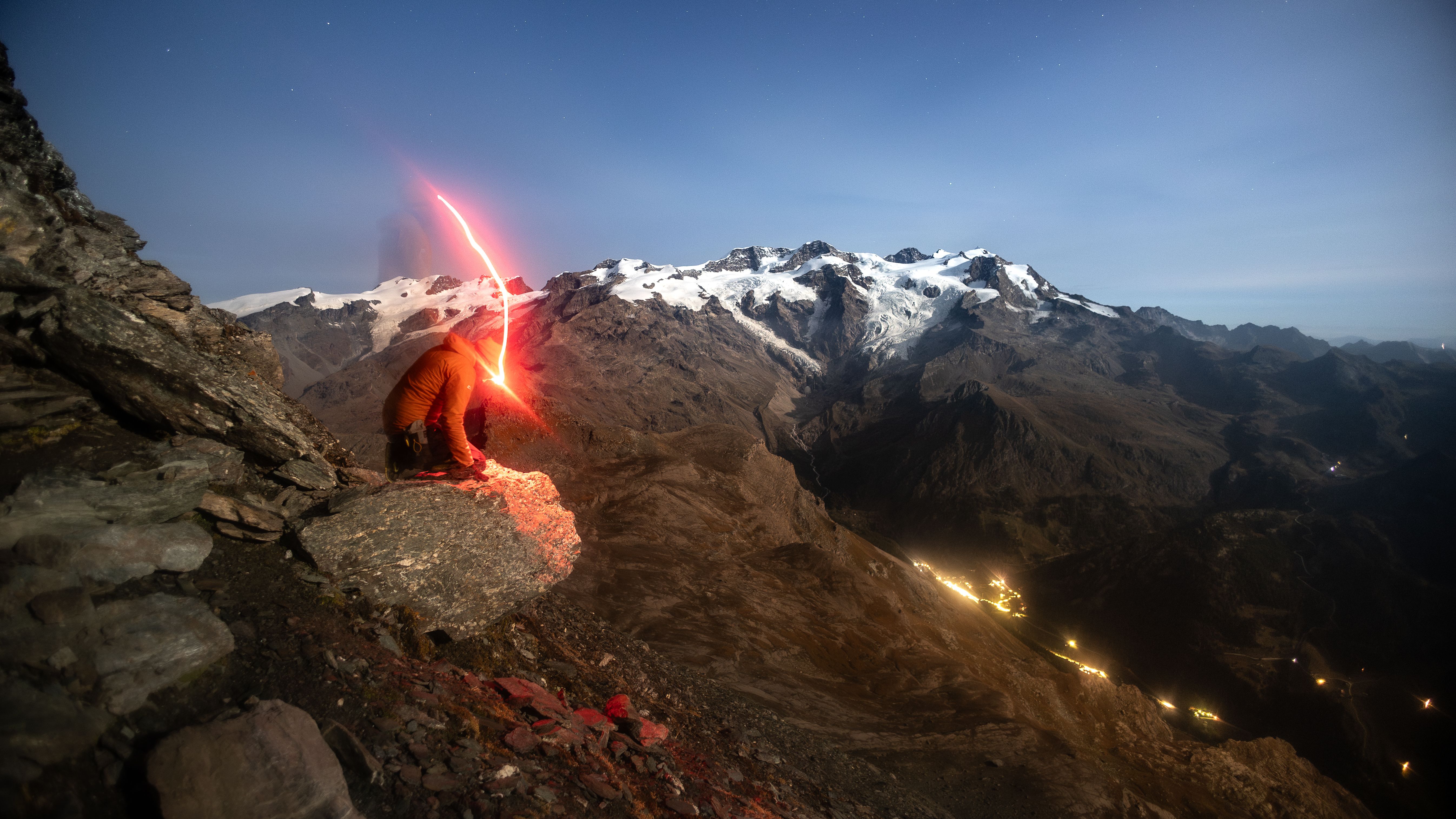 A solitary hiker with a torchlight kneeling on a rocky cliff, illuminating a vast alpine landscape under a night sky.