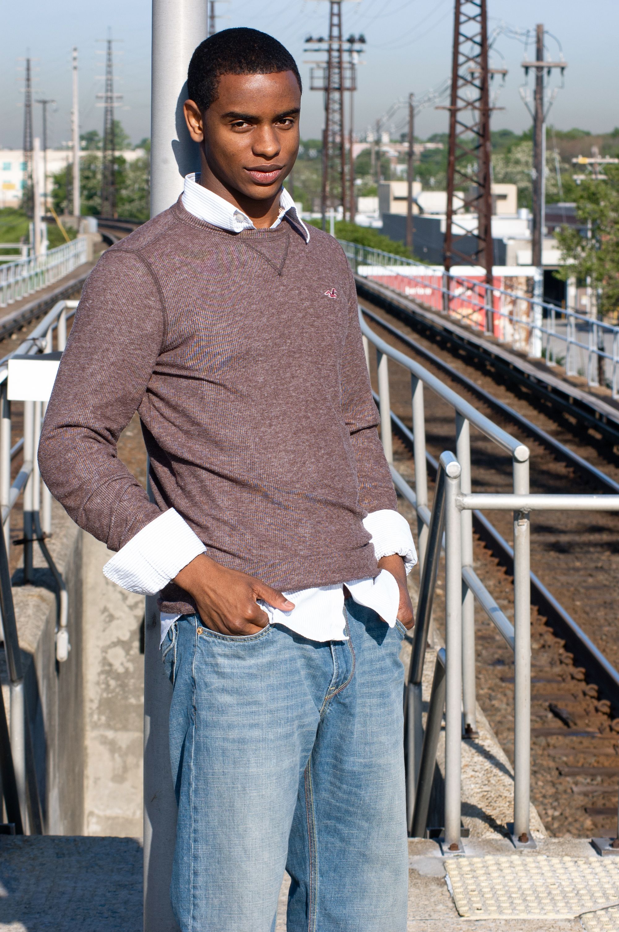 Young man posing in casual attire by railway tracks on a sunny day in West Hempstead.