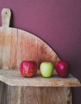 Three vibrant apples on a rustic wooden shelf against a textured wall, showcasing freshness and nutrition.
