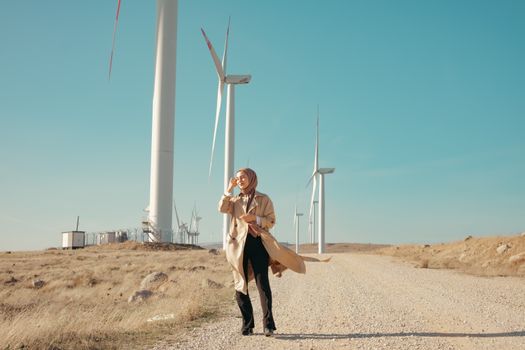 Woman in hijab on a dirt road near wind turbines, symbolizing sustainability and fashion.