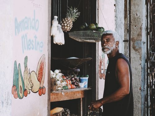 Free An elderly man stands by a fruit and vegetable stall at a city market. Vibrant produce on display. Stock Photo