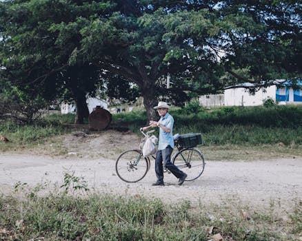 An elderly man walks his bike along a rural dirt road under a large tree.