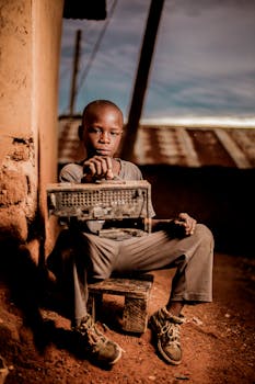 A young boy holding a vintage radio, seated outdoors against an earthy wall, captured during the day.