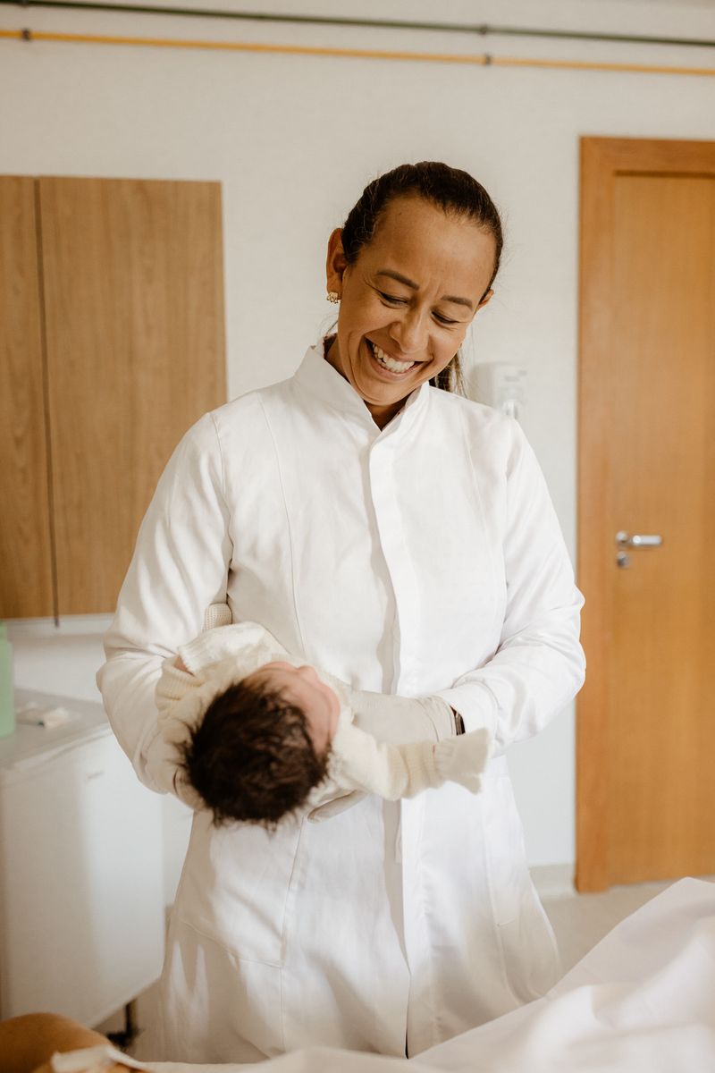 A Black woman midwife in a white coat smiles as she holds a newborn baby in a hospital ward.