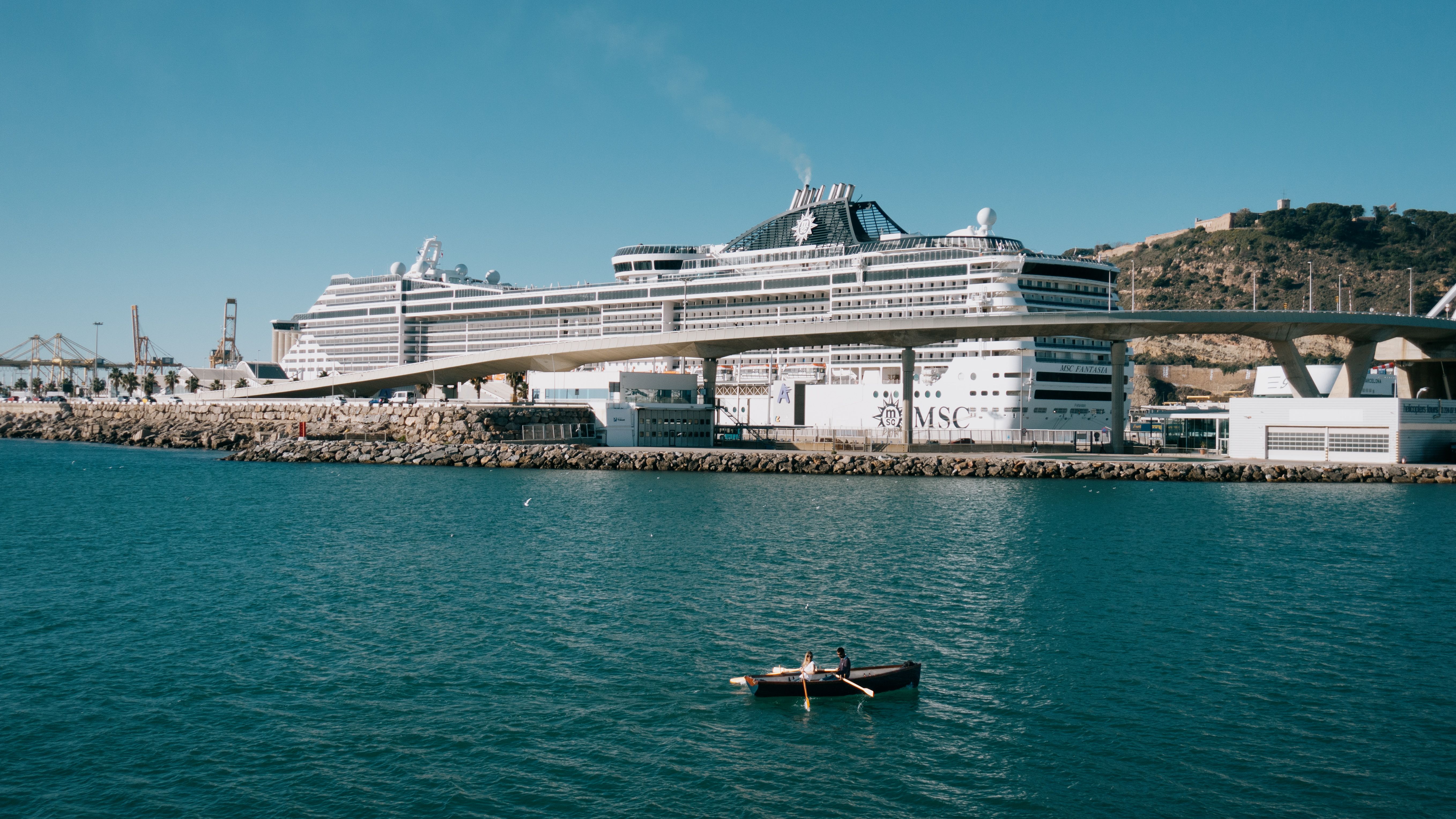 Large cruise ship docked at a sunny port with people rowing a boat in the foreground.
