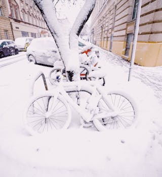 Snow blankets bicycles and cars on a quiet city street, creating a serene winter scene.