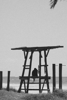 Black and white silhouette of a person sitting on a lifeguard tower by the coastline.