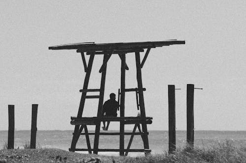 Black and White Photo of Man Sitting atop Wooden Lifeguard Tower