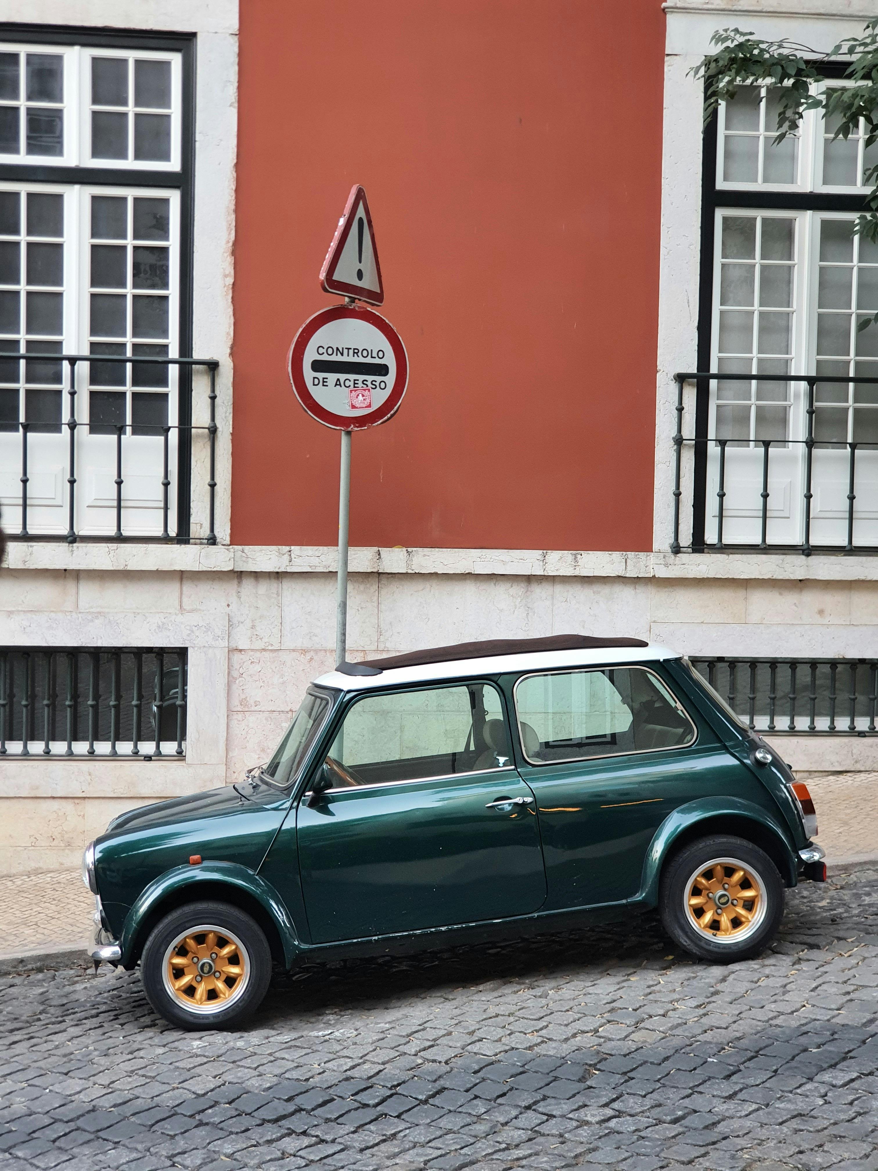 A green classic Mini Cooper is parked alongside a cobblestone street in Lisbon, Portugal.
