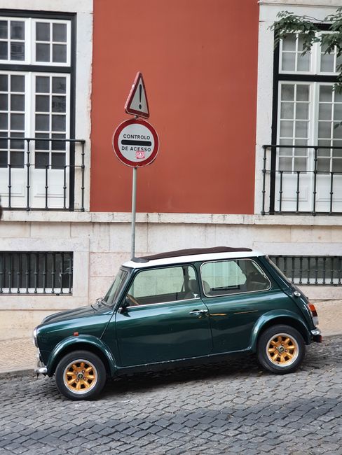 A green classic Mini Cooper is parked alongside a cobblestone street in Lisbon, Portugal.