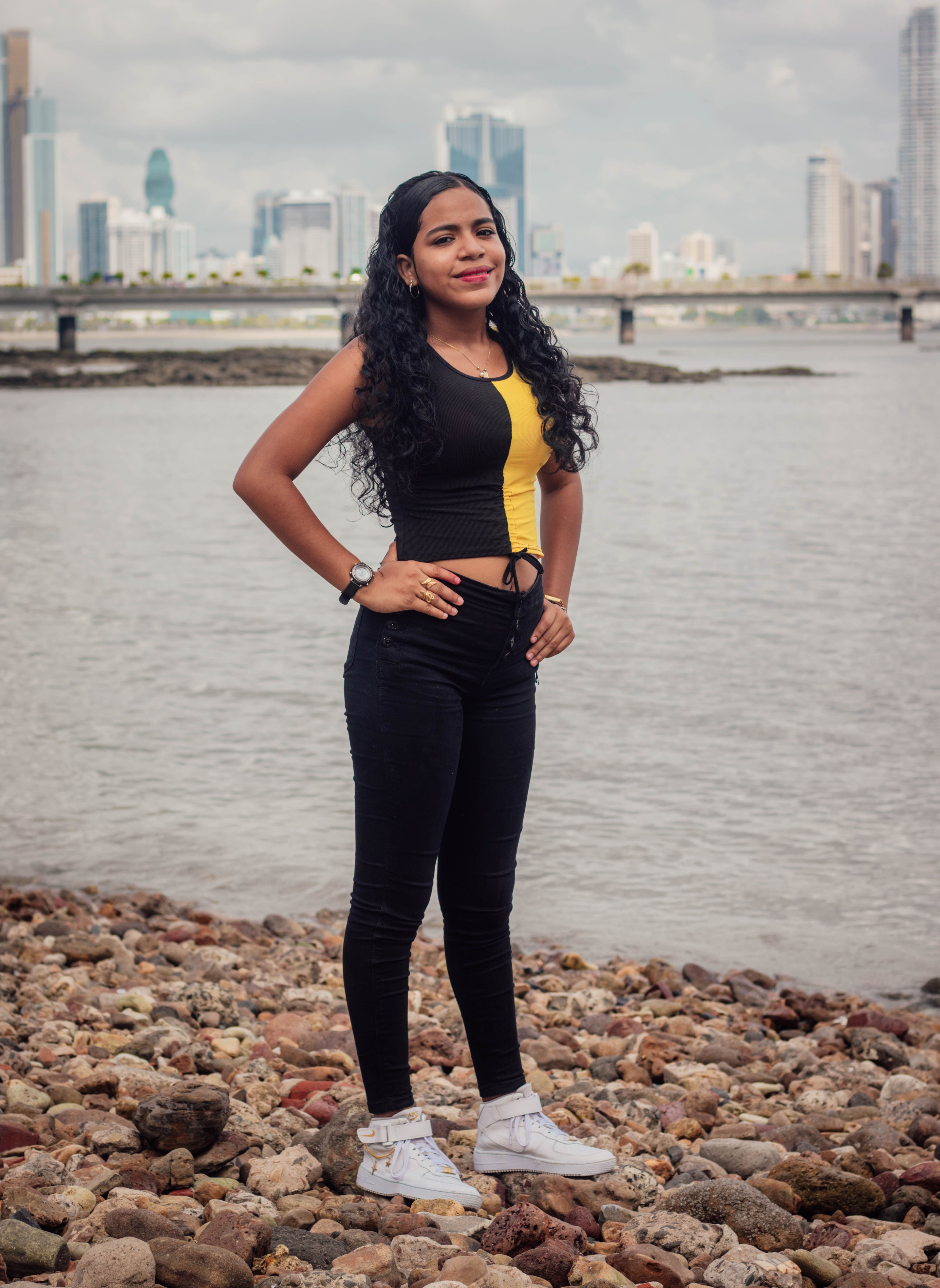 A confident young woman stands on rocky beach with city skyline in the background.