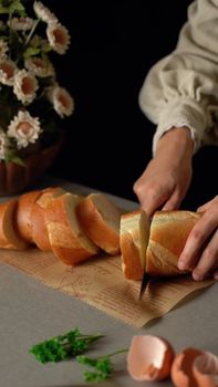 Close-up of hands slicing bread. Fresh ingredients and flowers in kitchen setting.