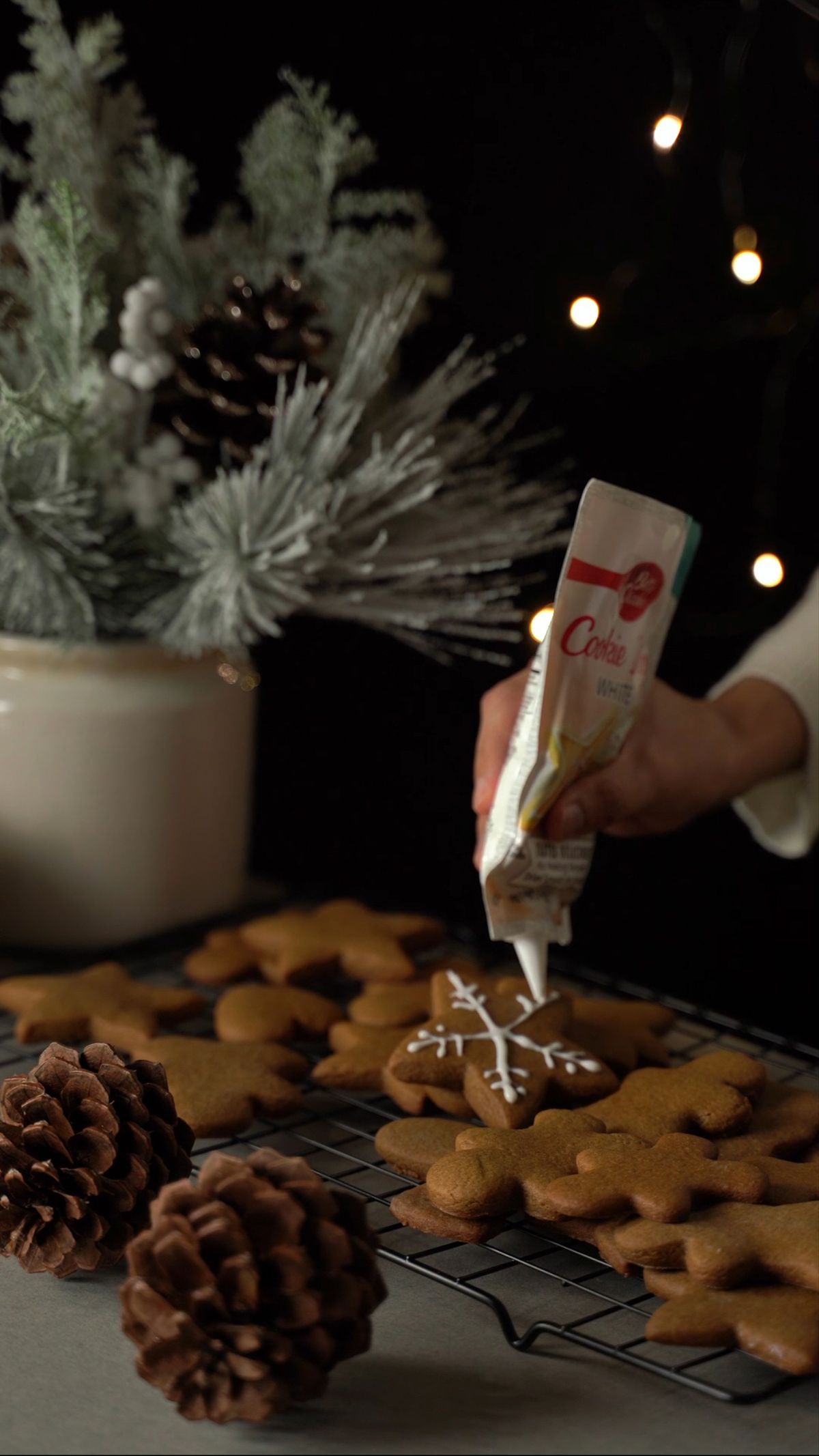 Hand piping white icing onto gingerbread and sugar cookies beside pine branches and pinecones