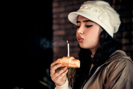 Portrait of a young woman with long hair blowing out a candle on a pastry indoors.
