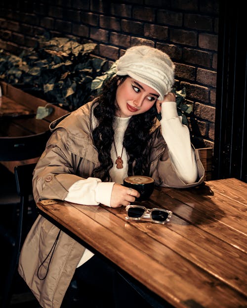 Model in a Beige Jacket over a White Sweater Sitting at the Table with Coffee