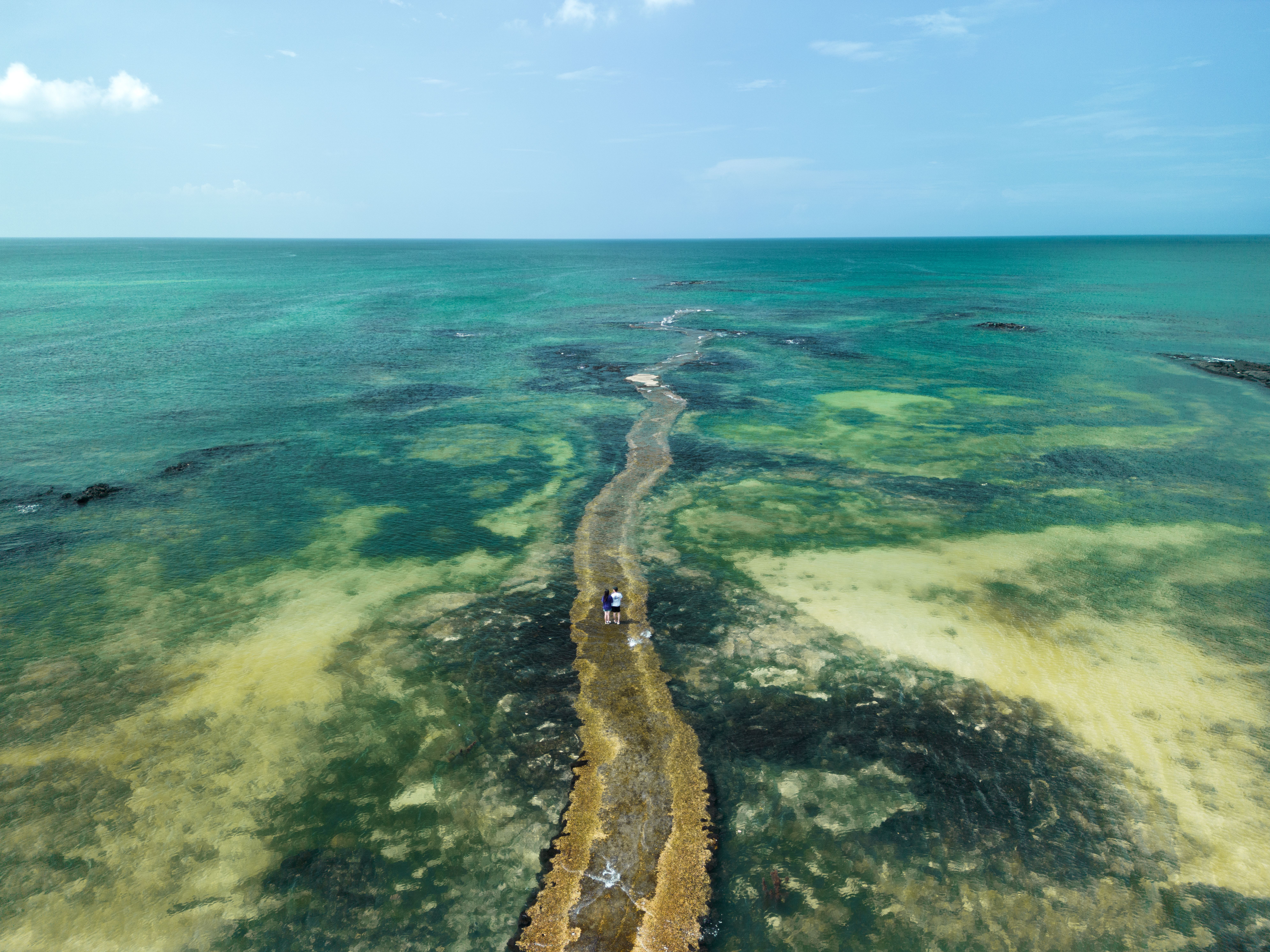 A stunning aerial view of a pathway extending into a clear tropical sea under a bright blue sky.