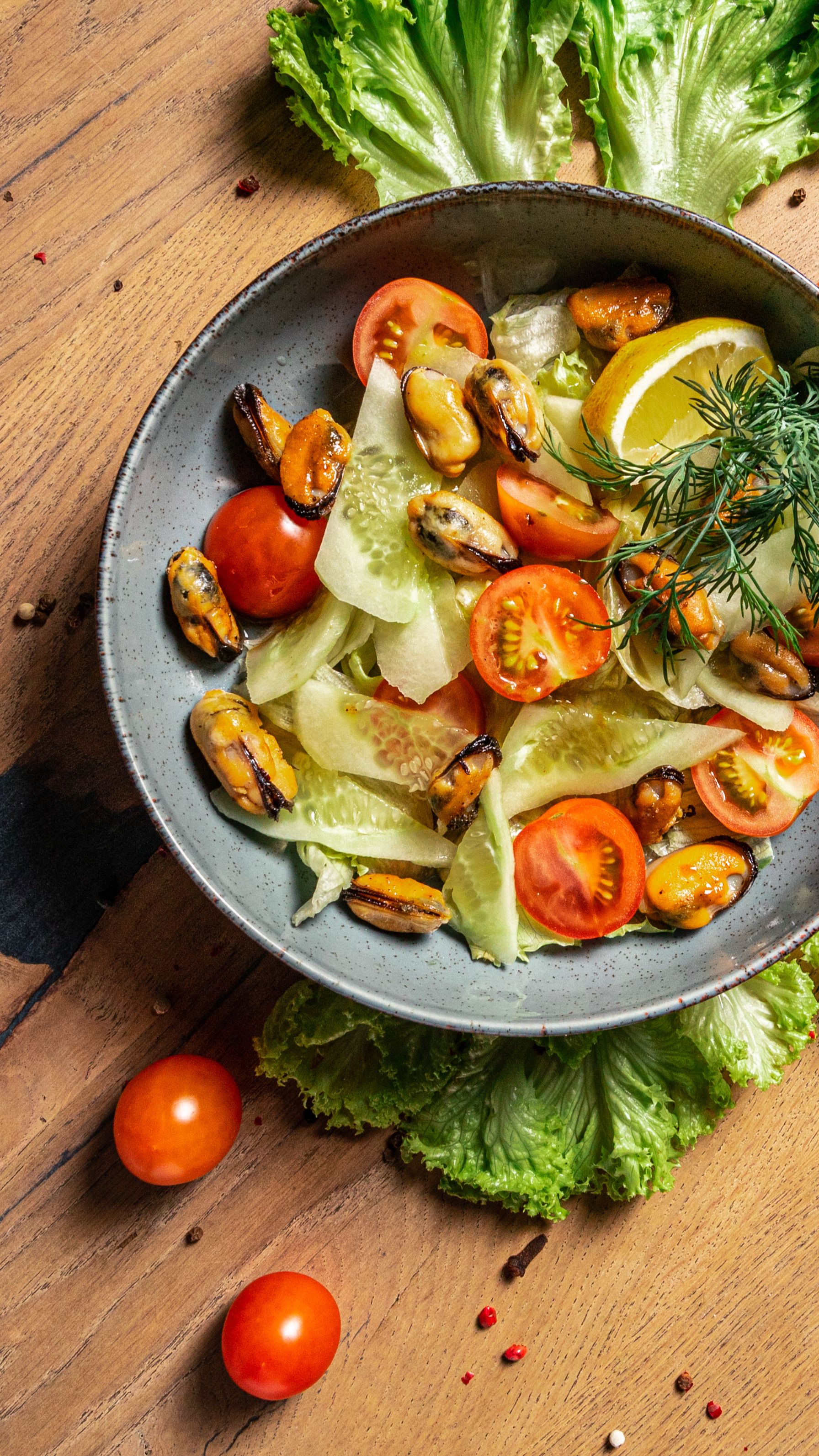 A vibrant bowl of salad with mussels, cherry tomatoes, cucumbers, and a lemon slice. Perfect for a refreshing meal.