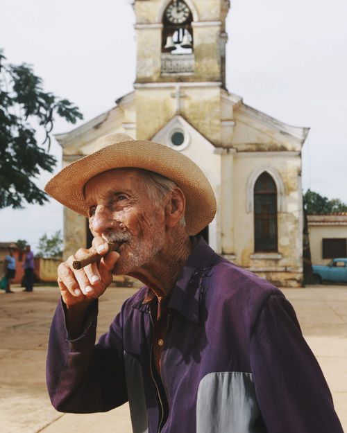 Free Portrait of an elderly man smoking a cigar in front of an old church. Rustic urban scene. Stock Photo