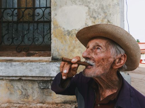 Free Portrait of an elderly man with a hat smoking a cigar against a weathered urban wall. Stock Photo