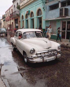 Vintage Chevrolet car driving through rainy city streets of Havana, Cuba, with colorful buildings and urban life.