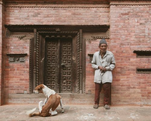 Free An elderly man stands near a vintage brick building adorned with intricate wooden carvings, accompanied by a sitting dog. Stock Photo