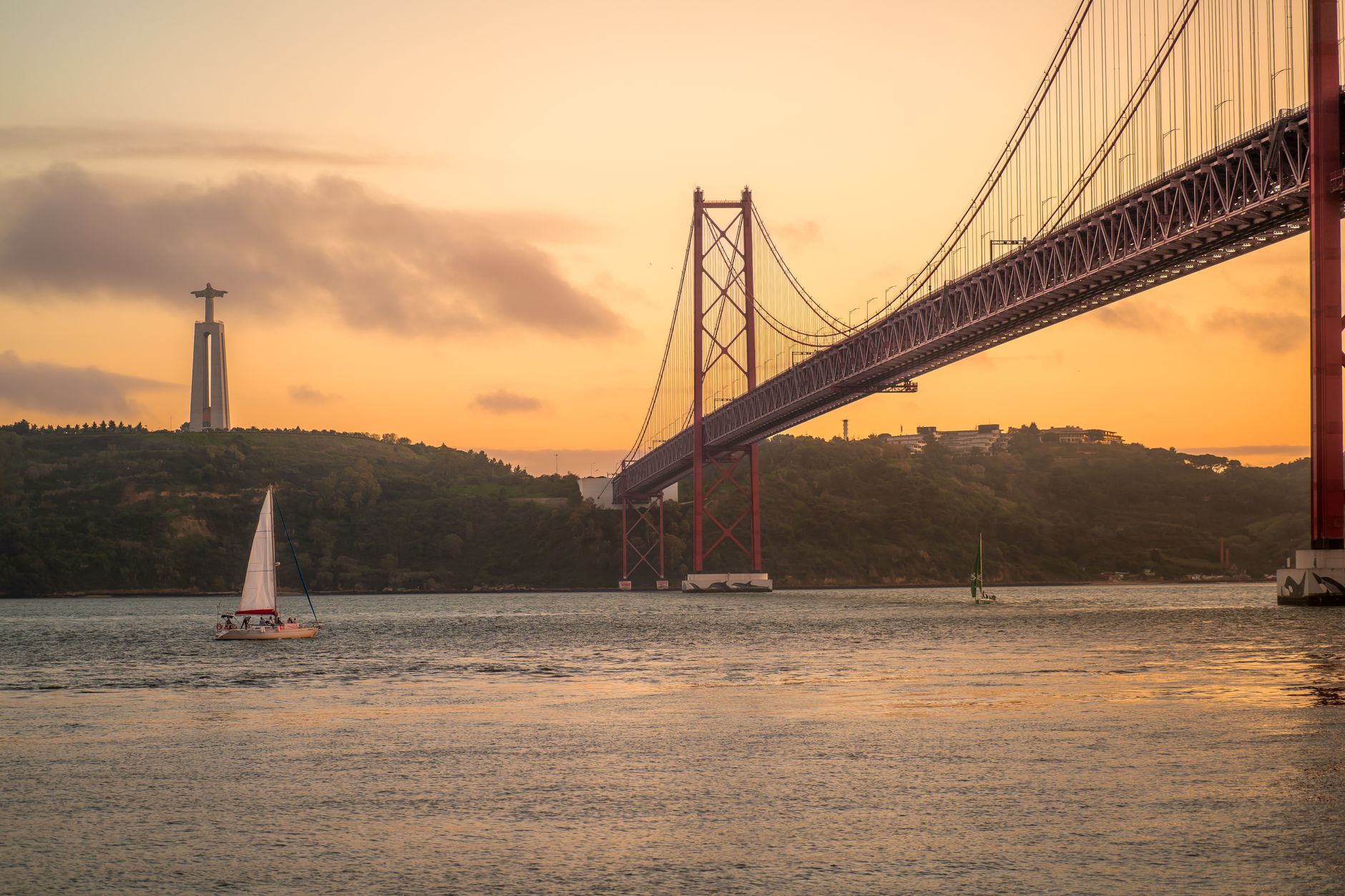 A serene view of a sailboat by the 25 de Abril Bridge at sunset in Lisbon, Portugal.