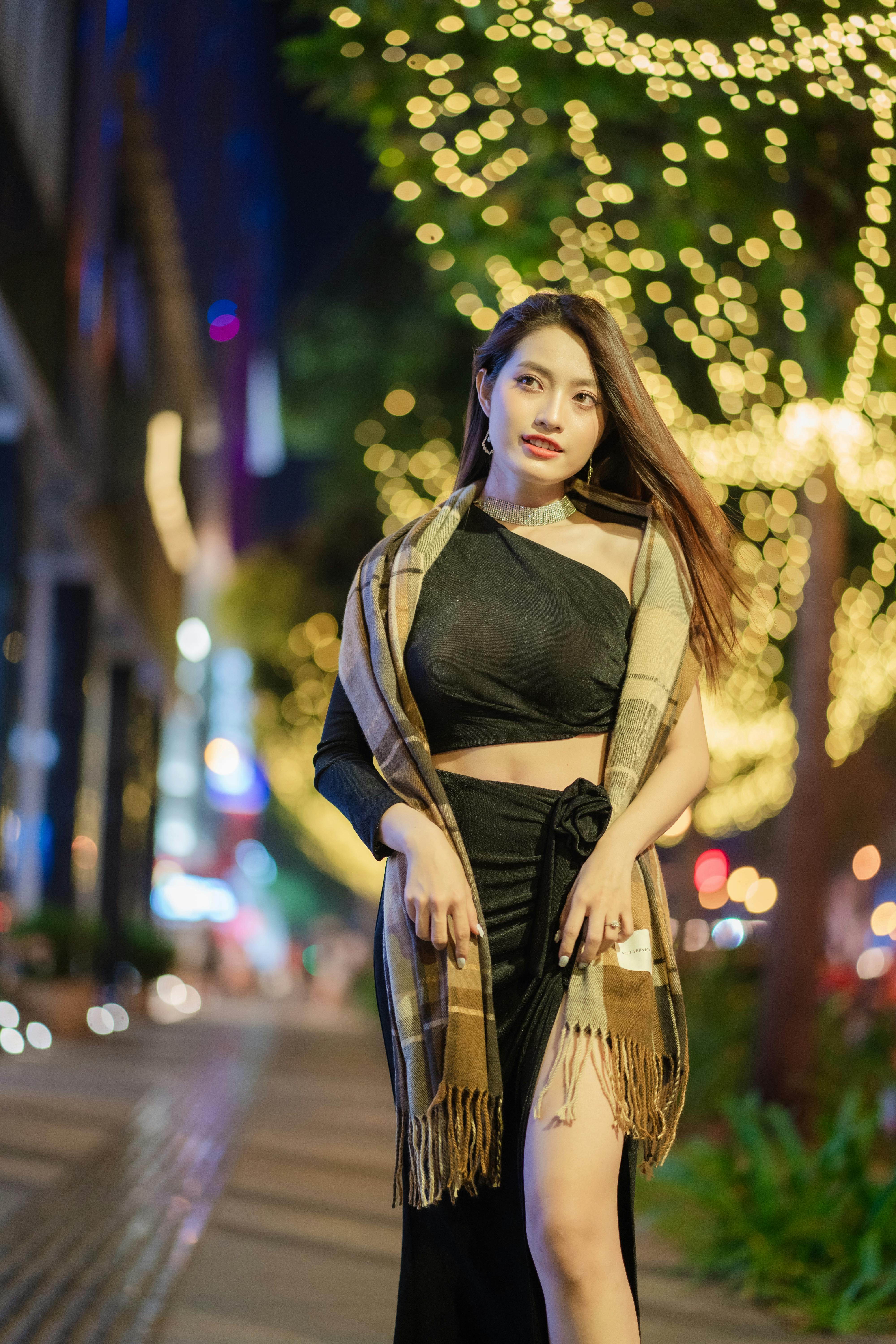 Elegant woman in a black dress and scarf posing on illuminated city street during night.