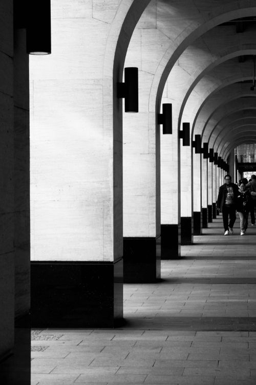 Free Monochrome image of an urban walkway with arches and pedestrians. Stock Photo