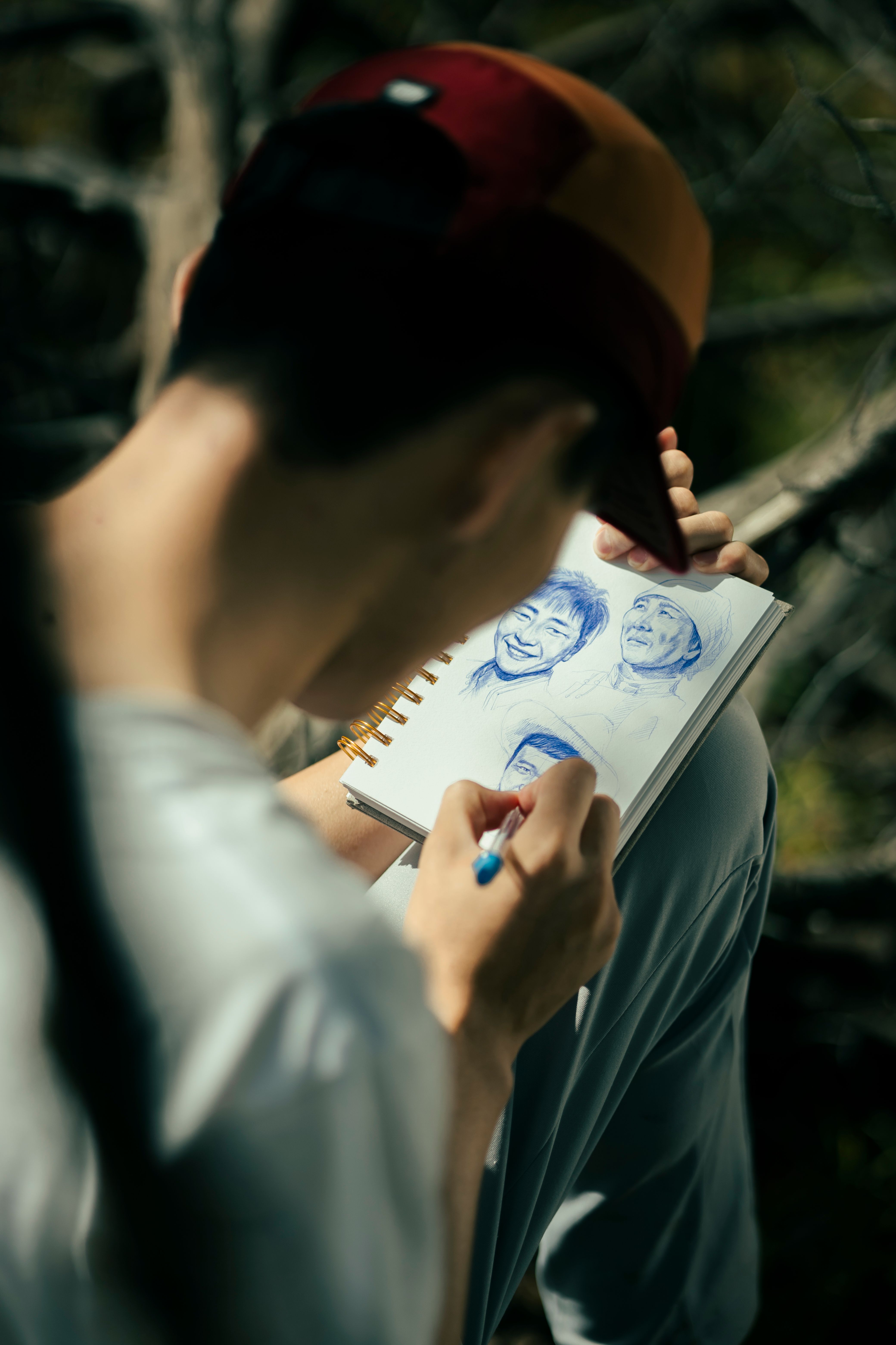 A man with a cap draws portraits in a notebook using a pencil, captured in daylight.