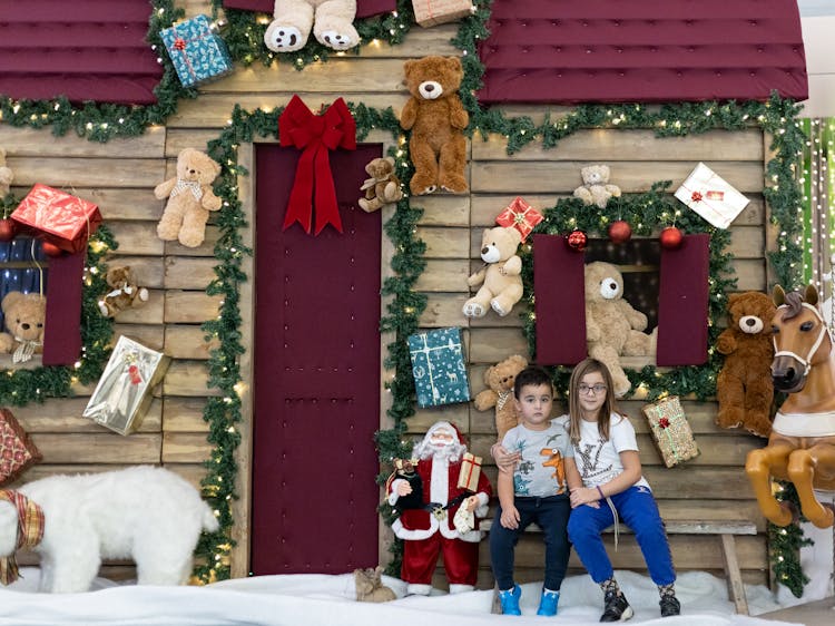 Children Sitting In Front Of A Christmas Hut Decorated With Toys 