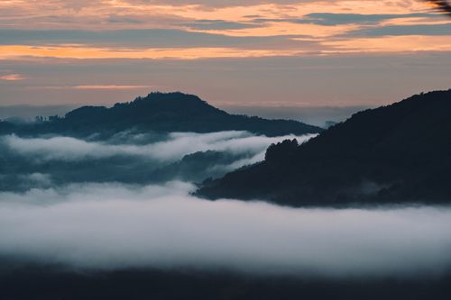 Free A tranquil dusk scene with mist enveloping mountain peaks under a colorful sky. Stock Photo