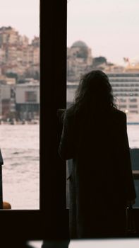 A woman in a coat gazes at a city skyline from indoors by a waterfront.