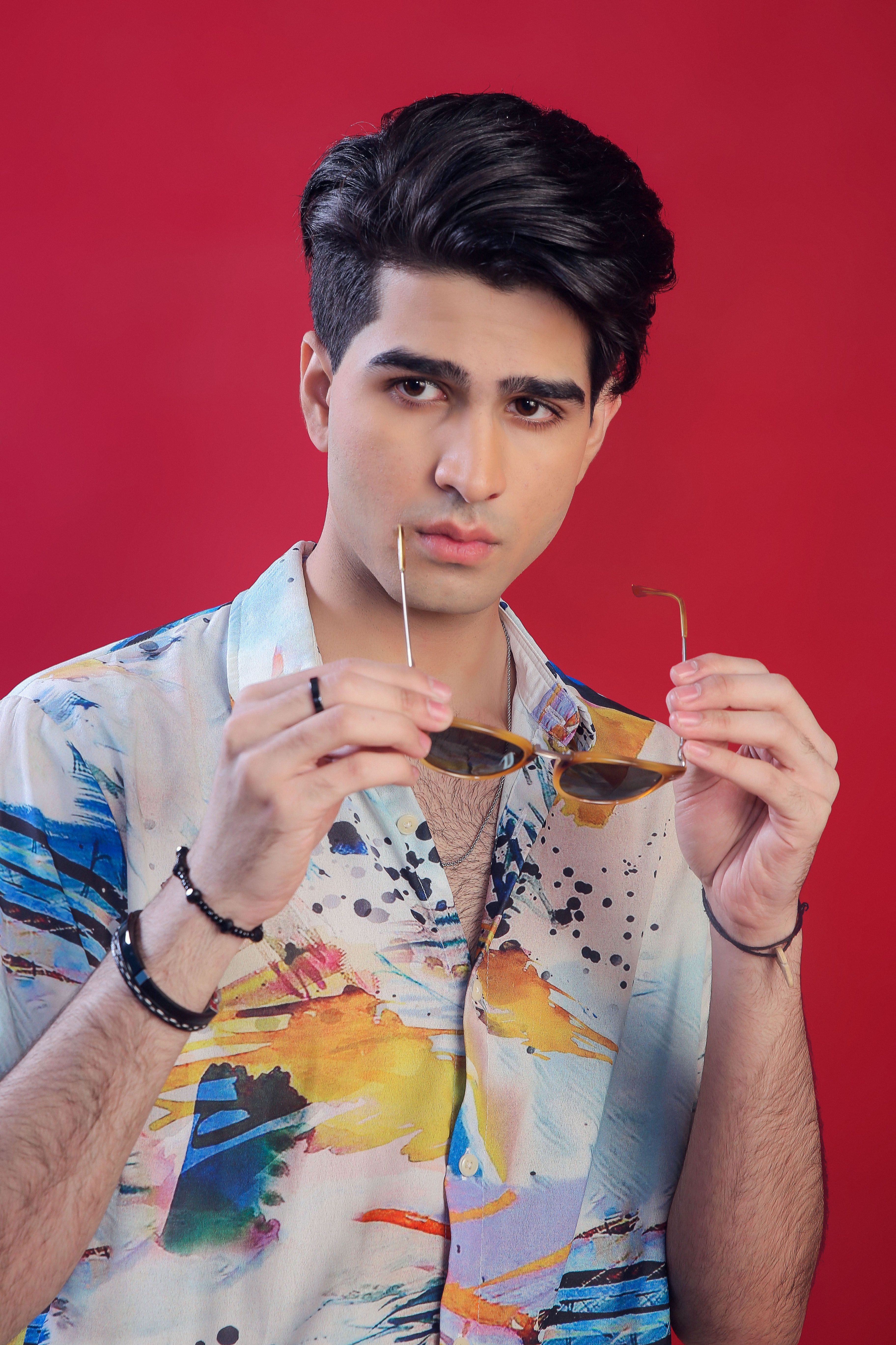 Stylish fashion portrait of a young man holding sunglasses against a red background.