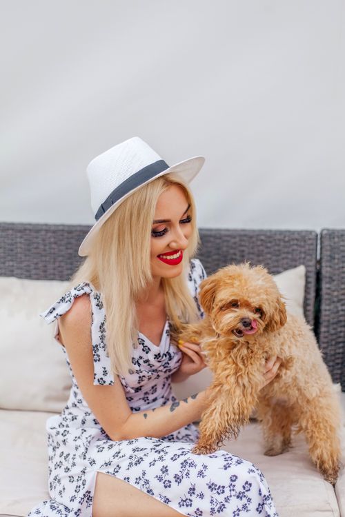 Free Smiling blonde woman in a hat, sitting outdoors with a fluffy dog, enjoying a sunny day. Stock Photo