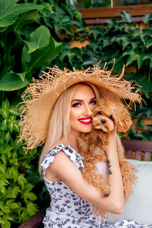 Free A stylish woman in a straw hat cuddles a poodle outdoors amidst lush greenery. Stock Photo
