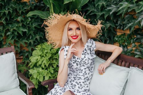 Free Blonde woman in summer dress with straw hat sitting on a bench, smiling. Stock Photo