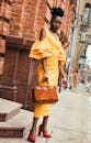 一位穿着黄色连衣裙的优雅女人，在城市街道上对着砖墙摆出姿势，展现着时尚与自信