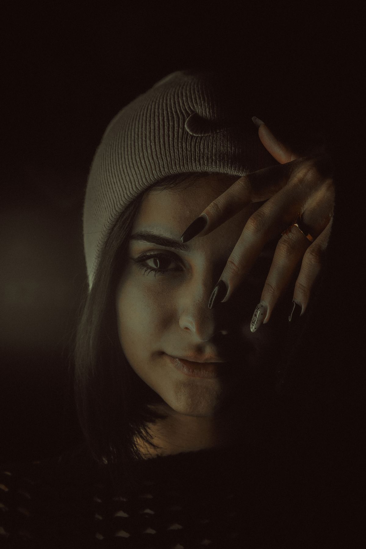 A dark, moody portrait of a young woman wearing a knit hat and showcasing embellished nail art.