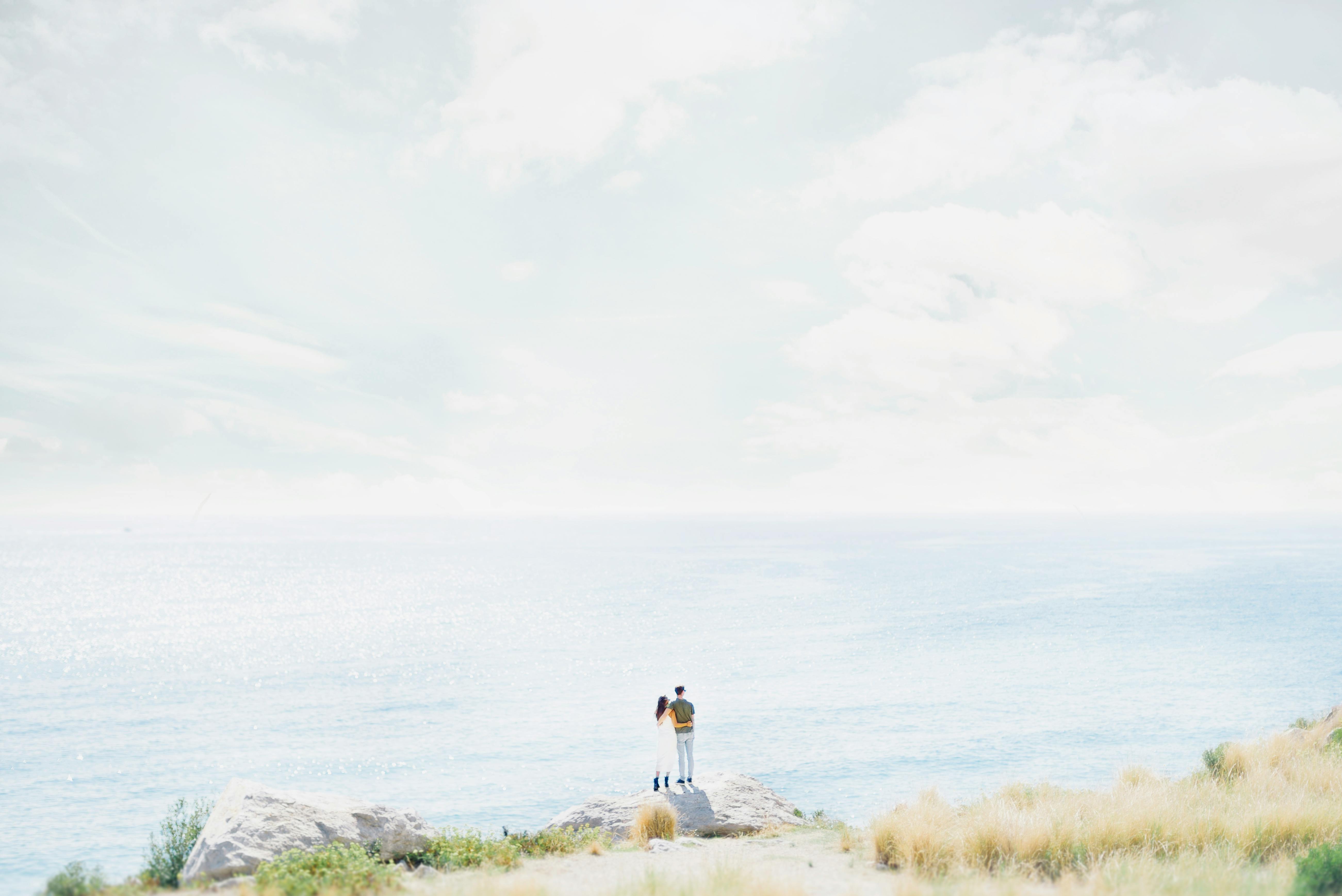 A romantic couple stands on a scenic seaside cliff, embracing the vast ocean view under a bright sky.