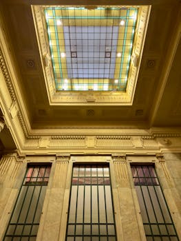 A beautifully ornamented interior with a stunning stained glass ceiling and large vertical windows.