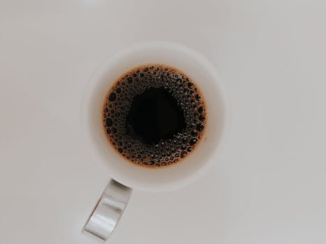 Minimalistic top view image of black coffee in a white cup with bubbles on its surface.